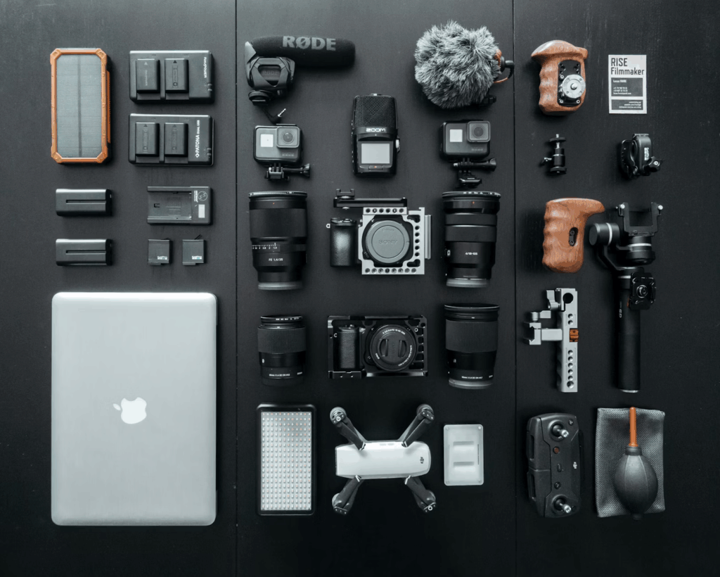 Flat lay photo of a MacBook surrounded by filmmaking gear, including cameras, lenses, microphones, drone, gimbal, and other accessories on a black background.
